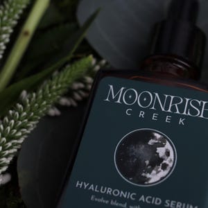 Op de afbeelding: Een donkere glazen fles Moonrise Creek Hyaluronzuur Serum met een zwarte druppelaar. Het etiket is teal met witte tekst en een maangrafiek. Witte en groene bladeren omringen de fles.
