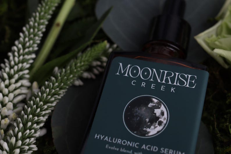 Op de afbeelding: Een donkere glazen fles Moonrise Creek Hyaluronzuur Serum met een zwarte druppelaar. Het etiket is teal met witte tekst en een maangrafiek. Witte en groene bladeren omringen de fles.