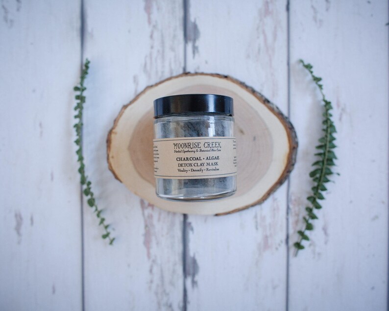 May include: A glass jar of Moonrise Creek Charcoal + Algae Detox Clay Mask on a wooden round slice. The jar has a black lid and a brown label with white text.