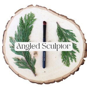 May include: A makeup brush with a black handle and an angled brown tip, featuring the text 'Moonrise Creek' on the handle. The brush is centered between two green cedar sprigs on a round, light-colored wood slice with the text 'Angled Sculptor'.