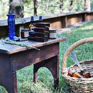 Peut inclure: Une table en bois rustique pr&eacute;sente des flacons d'apothicaire, des bocaux et un vase en verre bleu. Un panier en osier contient d'autres flacons et de la lavande. La sc&egrave;ne se d&eacute;roule en ext&eacute;rieur avec un arri&egrave;re-plan flou d'arbres et un banc en bois.