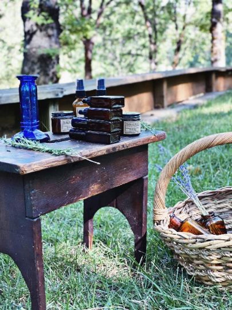 Peut inclure: Une table en bois rustique pr&eacute;sente des flacons d'apothicaire, des bocaux et un vase en verre bleu. Un panier en osier contient d'autres flacons et de la lavande. La sc&egrave;ne se d&eacute;roule en ext&eacute;rieur avec un arri&egrave;re-plan flou d'arbres et un banc en bois.