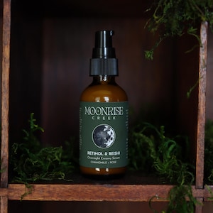 K&ouml;nnte beinhalten: Eine braune Glasflasche mit einem schwarzen Pumpspender. Das Etikett tr&auml;gt die Aufschrift "Moonrise Creek" und "Retinol & Reishi Overnight Creamy Serum". Die Flasche steht in einem Holzregal mit gr&uuml;nem Moos.