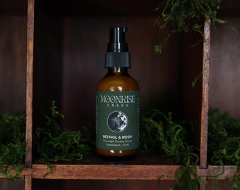 May include: A brown glass bottle with a black pump dispenser. The label reads "Moonrise Creek" and "Retinol & Reishi Overnight Creamy Serum". The bottle is set in a wooden shelf with green moss.