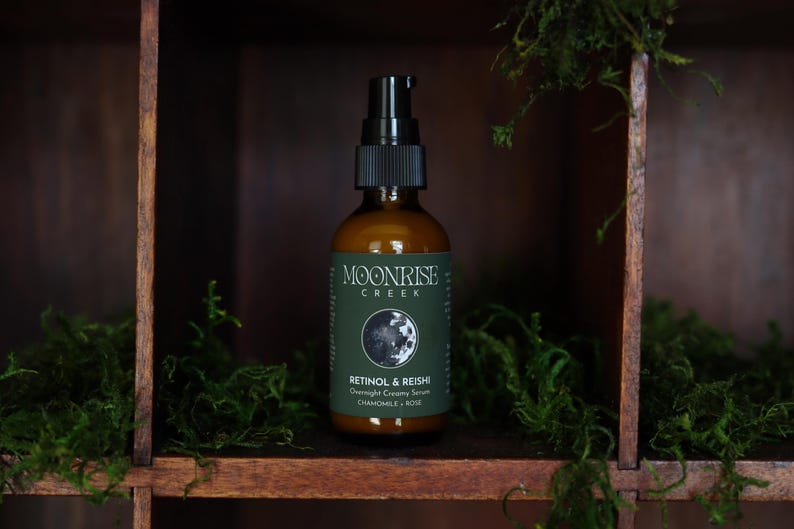 K&ouml;nnte beinhalten: Eine braune Glasflasche mit einem schwarzen Pumpspender. Das Etikett tr&auml;gt die Aufschrift "Moonrise Creek" und "Retinol & Reishi Overnight Creamy Serum". Die Flasche steht in einem Holzregal mit gr&uuml;nem Moos.