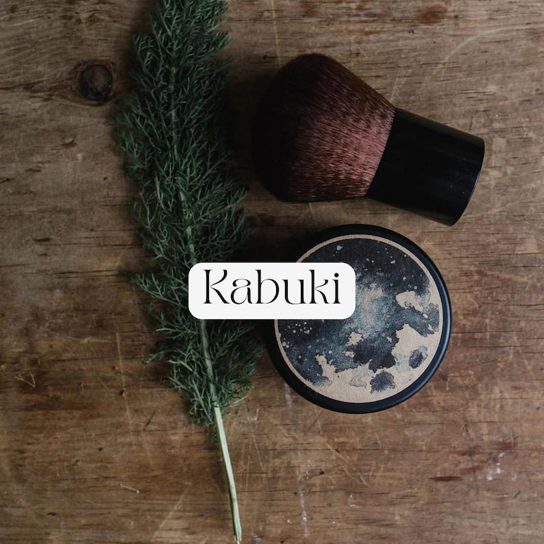 May include: A kabuki makeup brush with dark brown bristles and a black handle, positioned next to a round container with a dark, mottled blue and gold lid featuring the text Kabuki, alongside a green fern-like plant sprig.