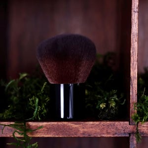 May include: A makeup brush with a dense, rounded head of dark brown bristles and a short, glossy black cylindrical handle. The brush is positioned upright, featuring soft, tapered fibers designed for powder application with a sleek, minimalist aesthetic.