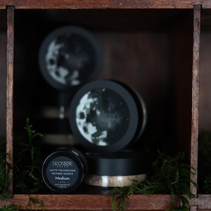 May include: A wooden display shelf holds several black containers of Moonrise Labs Matte Foundation Powder Sample. The containers have a black lid and a white label with the brand name and product details. The background features a moon design.