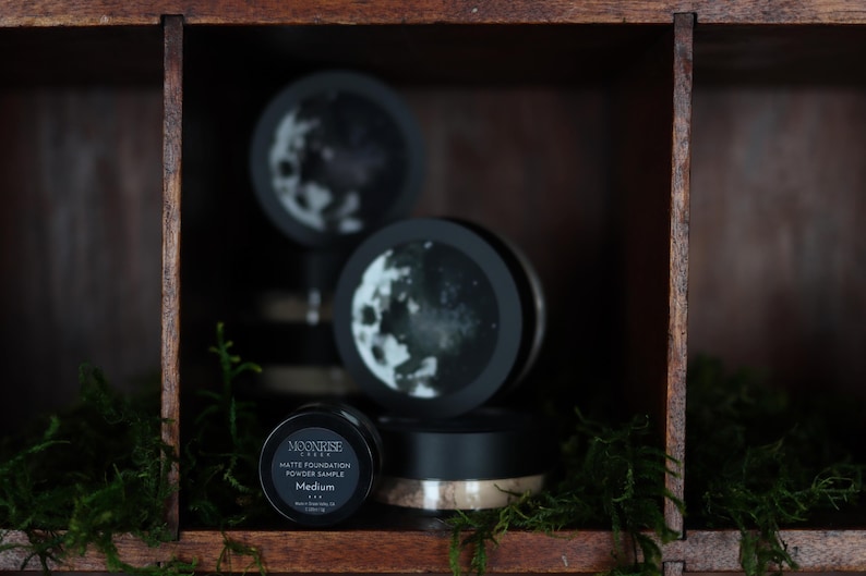 May include: A wooden display shelf holds several black containers of Moonrise Labs Matte Foundation Powder Sample. The containers have a black lid and a white label with the brand name and product details. The background features a moon design.