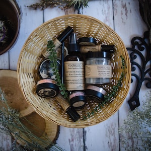 May include: A wicker basket filled with various skincare products, including a bottle of "Soft Rose Face & Body Mist" by Moonrise Craft, a jar of "Charcoal & Algae Detox Clay Mask" by Moonrise Craft, and several jars of face powder. The basket is on a white wooden surface with sprigs of greenery.
