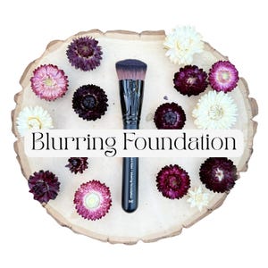 May include: A black foundation makeup brush with a flat top and dense bristles, labeled Blurring Foundation, surrounded by dried purple, pink, and white strawflowers on a light-colored wooden slice with a natural, irregular bark edge.