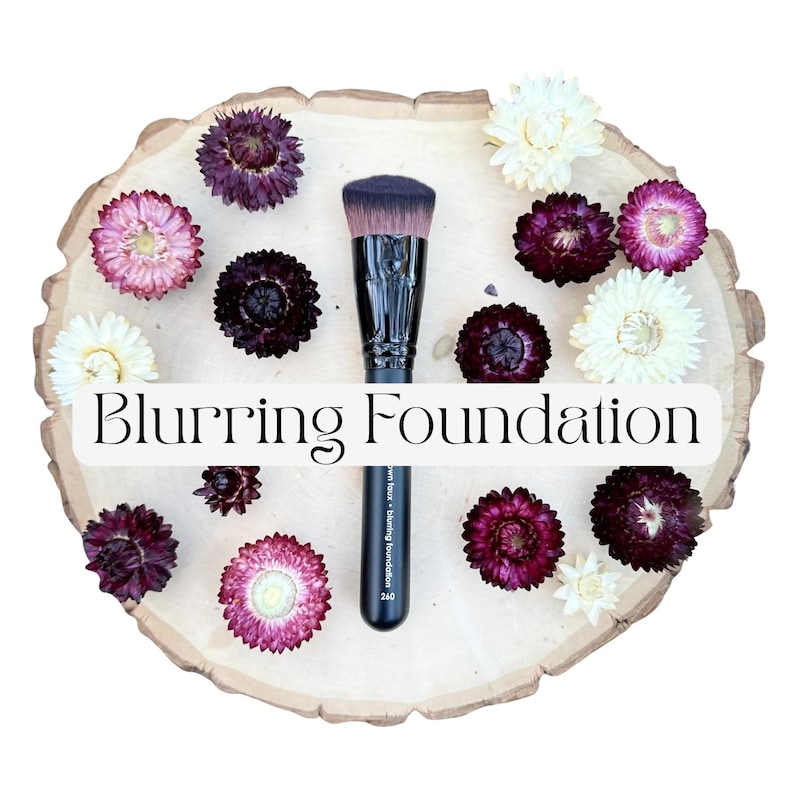 May include: A black foundation makeup brush with a flat top and dense bristles, labeled Blurring Foundation, surrounded by dried purple, pink, and white strawflowers on a light-colored wooden slice with a natural, irregular bark edge.