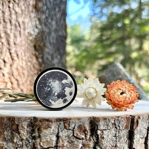 May include: A black and white moon print on a round wooden disc, with two dried flowers, one white and one orange, on a wooden log.