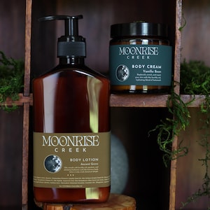 Może przedstawiać: Brązowa butelka balsamu do ciała i słoik kremu do ciała Moonrise Creek. Butelka balsamu ma czarną pompkę i etykietę z napisem "Body Lotion Ancient Grove". Słoik kremu do ciała jest oznaczony napisem "Body Cream Vanilla Bean".