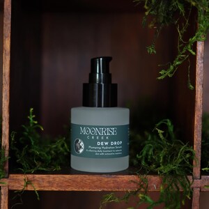 May include: A frosted glass bottle with a black pump dispenser, labeled "Moonrise Creek Dew Drop Plumping Hydration Serum." The bottle sits on a wooden shelf, surrounded by green moss. The background is a dark wooden structure.