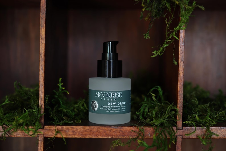 May include: A frosted glass bottle with a black pump dispenser, labeled "Moonrise Creek Dew Drop Plumping Hydration Serum." The bottle sits on a wooden shelf, surrounded by green moss. The background is a dark wooden structure.
