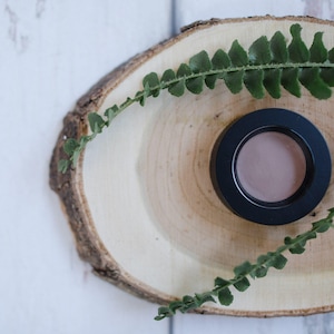 May include: A round, black container with a lid, containing a light brown cream or powder, sits on a wooden slice with green leaves.