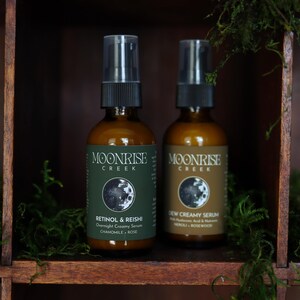 K&ouml;nnte beinhalten: Zwei braune Glasflaschen mit Moonrise Creek Hautpflegeprodukten. Eine Flasche ist mit "Retinol & Reishi Overnight Creamy Serum" beschriftet, die andere mit "New Creamy Serum". Beide haben schwarze Spr&uuml;hk&ouml;pfe und stehen auf einem Holzregal mit Gr&uuml;n.