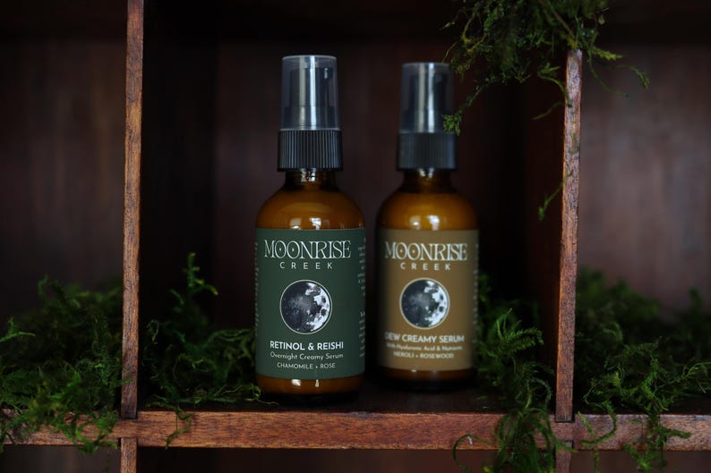 K&ouml;nnte beinhalten: Zwei braune Glasflaschen mit Moonrise Creek Hautpflegeprodukten. Eine Flasche ist mit "Retinol & Reishi Overnight Creamy Serum" beschriftet, die andere mit "New Creamy Serum". Beide haben schwarze Spr&uuml;hk&ouml;pfe und stehen auf einem Holzregal mit Gr&uuml;n.