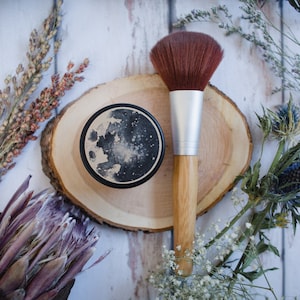 May include: A make-up brush with a brown brush head and a silver ferrule rests on a wooden slice next to a compact with a moon design. Dried flowers and foliage surround the beauty products, creating a natural aesthetic.