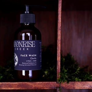 May include: A dark amber glass bottle with a black pump dispenser containing face wash. The label reads Moonrise Creek, Face Wash, Clarify, Citrus + Herb, Botanical Exfoliation & Detoxing Activated Charcoal, with a circular graphic of a moon.