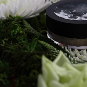 May include: A black jar with a silver rim and a dark lid, possibly containing a cosmetic product, rests amidst green moss, white flowers, and greenery. The lid has a design on it. The image has a soft focus.