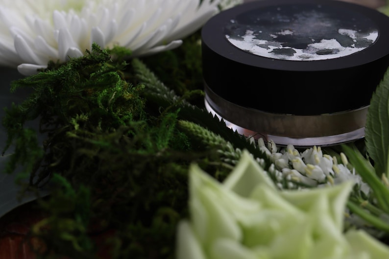May include: A black jar with a silver rim and a dark lid, possibly containing a cosmetic product, rests amidst green moss, white flowers, and greenery. The lid has a design on it. The image has a soft focus.