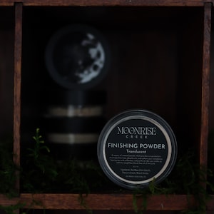 May include: A black container of Moonrise Creek Finishing Powder, translucent, is displayed on a wooden shelf. The container has a white label with black text. The shelf is part of a dark wooden cabinet, with green moss and other containers in the background.