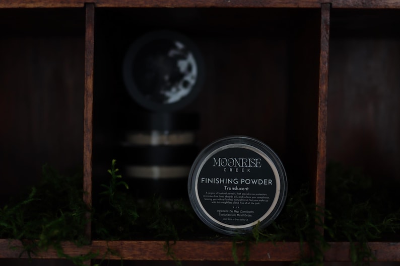 May include: A black container of Moonrise Creek Finishing Powder, translucent, is displayed on a wooden shelf. The container has a white label with black text. The shelf is part of a dark wooden cabinet, with green moss and other containers in the background.