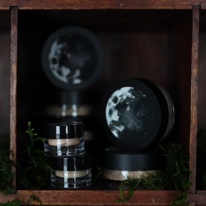 May include: Cosmetic jars and containers are arranged on a wooden shelf. The clear jars with black lids are stacked and paired with moon-themed decorative toppers. Green moss provides a natural accent to the display.