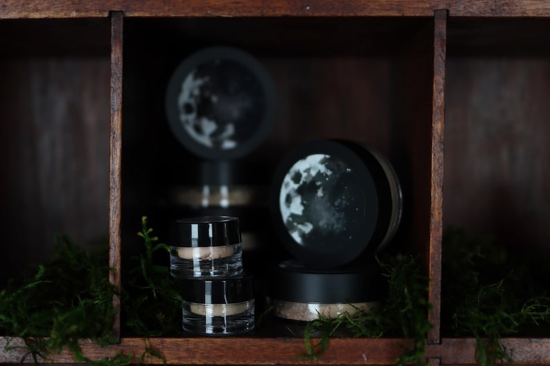 May include: Cosmetic jars and containers are arranged on a wooden shelf. The clear jars with black lids are stacked and paired with moon-themed decorative toppers. Green moss provides a natural accent to the display.
