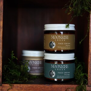 Puede incluir: Tres tarros de cristal de crema facial Moonrise Creek apilados en una estanter&iacute;a de madera. Los tarros tienen tapas blancas y etiquetas marrones con el nombre de la marca y los detalles del producto. La estanter&iacute;a est&aacute; rodeada de musgo verde, creando una est&eacute;tica natural y terrosa.