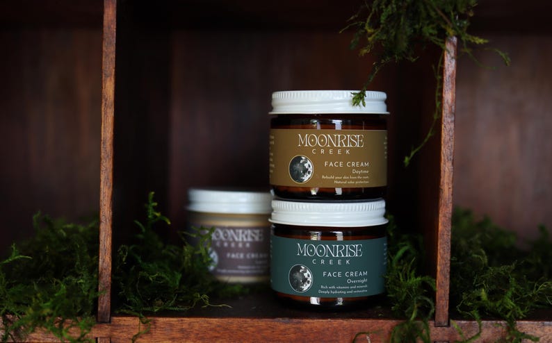 Puede incluir: Tres tarros de cristal de crema facial Moonrise Creek apilados en una estanter&iacute;a de madera. Los tarros tienen tapas blancas y etiquetas marrones con el nombre de la marca y los detalles del producto. La estanter&iacute;a est&aacute; rodeada de musgo verde, creando una est&eacute;tica natural y terrosa.