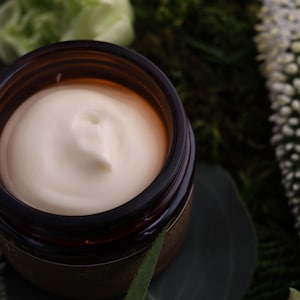 Puede incluir: Un tarro de crema de color beige claro, sobre un fondo de vegetaci&oacute;n y flores blancas. La crema tiene una textura suave y arremolinada, y el tarro es de color &aacute;mbar oscuro.