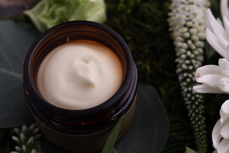 Puede incluir: Un tarro de crema de color beige claro, sobre un fondo de vegetaci&oacute;n y flores blancas. La crema tiene una textura suave y arremolinada, y el tarro es de color &aacute;mbar oscuro.