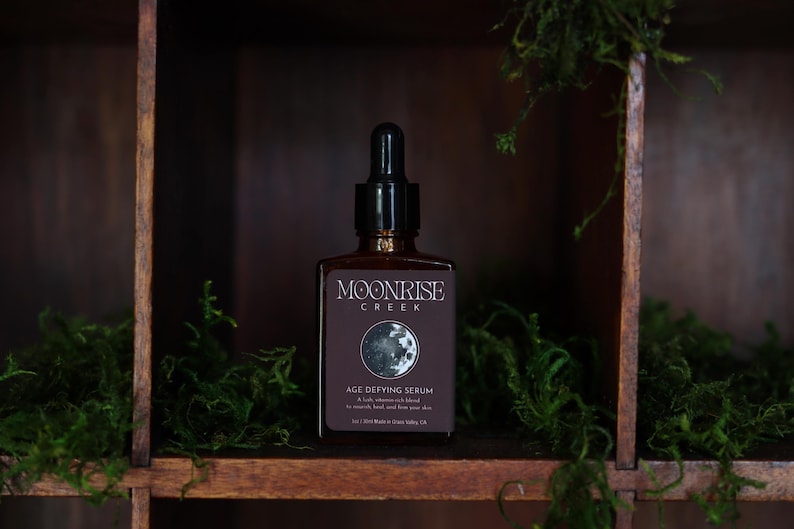 May include: A brown glass bottle of Moonrise Creek Age Defying Serum with a black dropper. The label features the brand name, a moon graphic, and product details. The bottle is displayed on a wooden shelf with green moss.