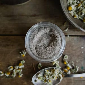 May include: A glass jar filled with a gray powder, surrounded by dried chamomile flowers on a wooden surface.