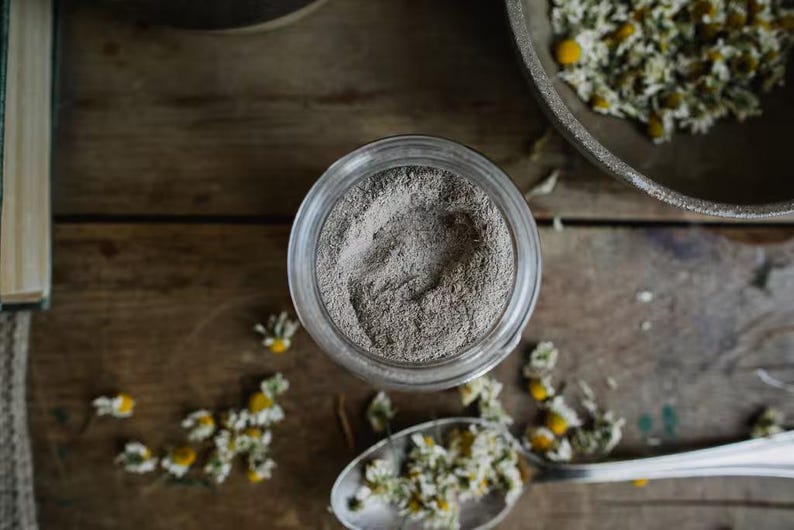 May include: A glass jar filled with a gray powder, surrounded by dried chamomile flowers on a wooden surface.