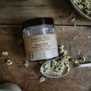 May include: A glass jar with a black lid containing a light gray powder. The label on the jar reads "Moonrise Creek Herbal Apothecary & Botanical Skin Care Flower Child Moroccan Clay Mask Cleanse - Repair - Nourish".
