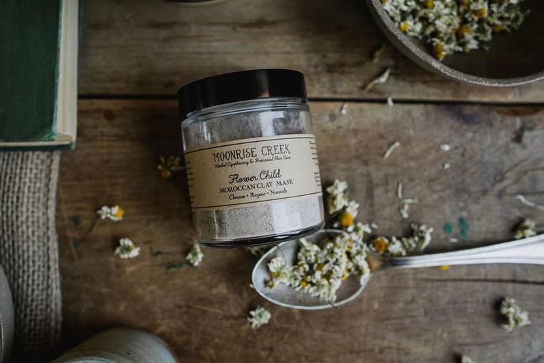 May include: A glass jar with a black lid containing a light gray powder. The label on the jar reads "Moonrise Creek Herbal Apothecary & Botanical Skin Care Flower Child Moroccan Clay Mask Cleanse - Repair - Nourish".