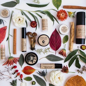 Peut inclure: Vue a&eacute;rienne de produits de beaut&eacute; naturels, dont des baumes &agrave; l&egrave;vres, des cr&egrave;mes pour le visage et un flacon de fond de teint, dispos&eacute;s avec des feuilles, des fleurs et des b&acirc;tons de cannelle. Les produits sont &eacute;tiquet&eacute;s "Moonrise Creek". La palette de couleurs est terreuse, avec des rouges, des verts et des bruns.