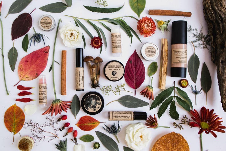 Peut inclure: Vue a&eacute;rienne de produits de beaut&eacute; naturels, dont des baumes &agrave; l&egrave;vres, des cr&egrave;mes pour le visage et un flacon de fond de teint, dispos&eacute;s avec des feuilles, des fleurs et des b&acirc;tons de cannelle. Les produits sont &eacute;tiquet&eacute;s "Moonrise Creek". La palette de couleurs est terreuse, avec des rouges, des verts et des bruns.