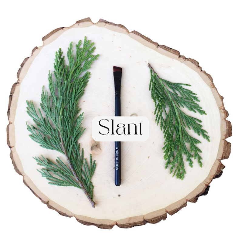 May include: A black makeup brush with an angled tip, featuring the text 'Slant' and 'MOONRISE CREEK' printed on the handle. The brush is positioned vertically between two green evergreen sprigs on a light-colored, circular wooden slice.