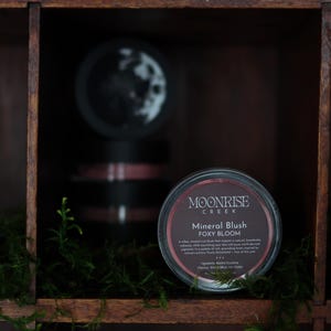 Può includere: Una mensola in legno scuro espone il fard minerale Moonrise Creek Foxy Bloom. Il fard è in un contenitore rotondo con bordo argentato ed etichetta scura. Muschio verde e piccole piante in primo piano.
