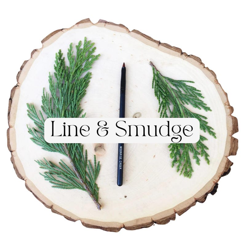 May include: A black cosmetic pencil with the text Moonrise Cheek printed on the side, positioned vertically between two green cedar sprigs on a round, light-colored wooden slice. A white rectangular overlay displays the text Line & Smudge.