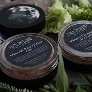 May include: Three pots of Moonrise Creek Mineral Foundation, set amongst greenery and white flowers. The containers have black lids with labels reading "Mineral Foundation" and "Fair" or "Matte Fair". One lid displays a moon image.