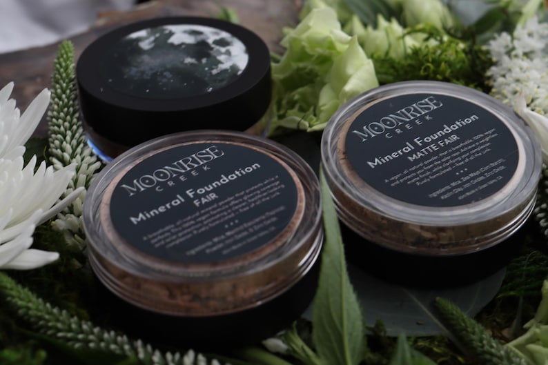May include: Three pots of Moonrise Creek Mineral Foundation, set amongst greenery and white flowers. The containers have black lids with labels reading "Mineral Foundation" and "Fair" or "Matte Fair". One lid displays a moon image.