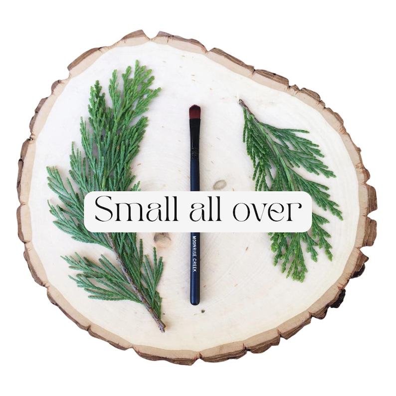 May include: A black makeup brush with a dark red tip and the text MOONRISE CREEK printed on the handle. The brush is positioned vertically between two green cedar sprigs on a light-colored wooden slice with the text Small all over in the center.
