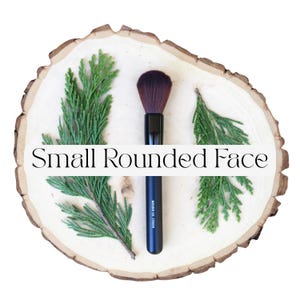 May include: A makeup brush with a black handle and dark brown bristles, positioned vertically between two green cedar sprigs on a light-colored wood slice. The black handle features the white text Moonrise Creek and Small Rounded Face.