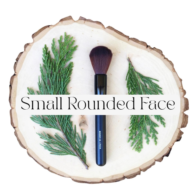 May include: A makeup brush with a black handle and dark brown bristles, positioned vertically between two green cedar sprigs on a light-colored wood slice. The black handle features the white text Moonrise Creek and Small Rounded Face.